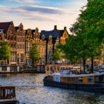 Scenic view of Amsterdam canal houses and boats at sunset with vibrant green trees.
